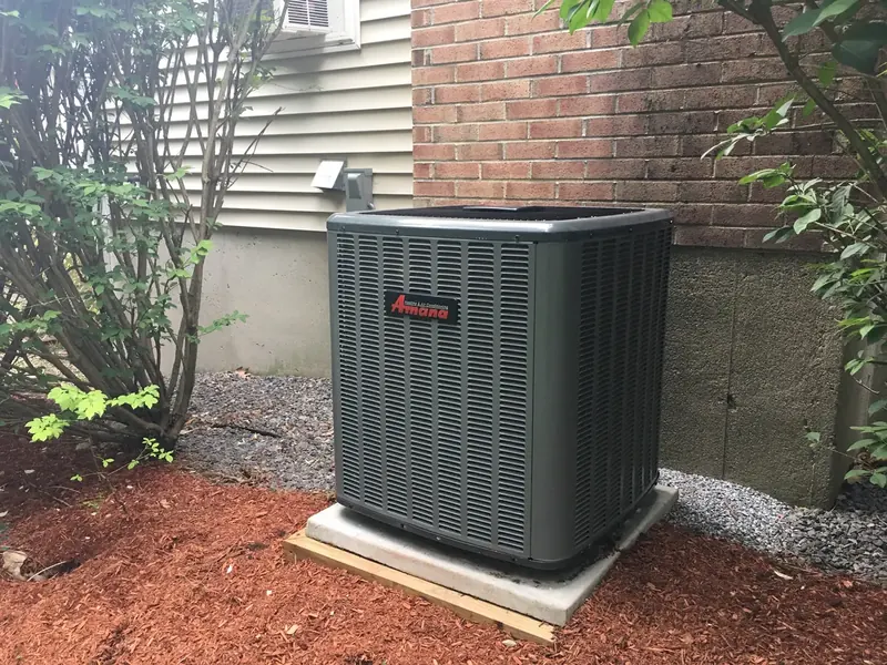 Amana central air conditioner condenser installed in a side yard beside a brick home by A.J. LeBlanc Heating in Southern NH