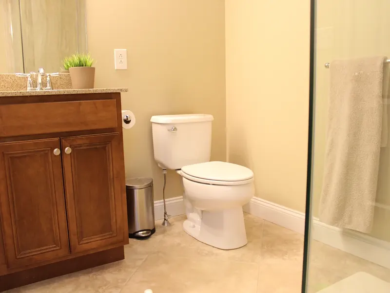 Bathroom with a centered two-piece toilet beside a brown wood vanity with granite counter, glass shower divider, and beige tile floor installed by A.J. LeBlanc Heating in a Southern NH home