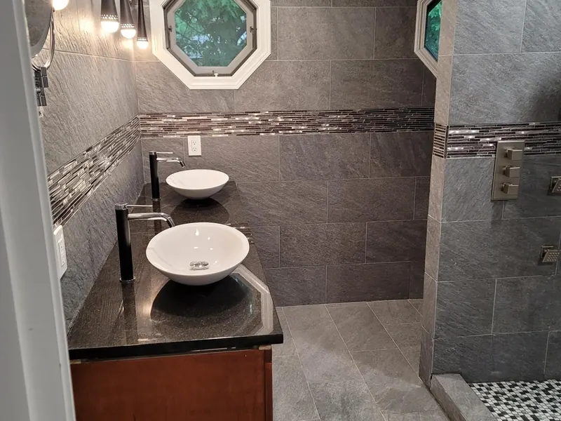 Two chrome wall-mount faucets above white vessel sinks on a wood vanity with black countertop, gray porcelain tile walls, glass mosaic accent band, and stained-glass octagonal window installed by A.J. LeBlanc Heating in a Southern NH home