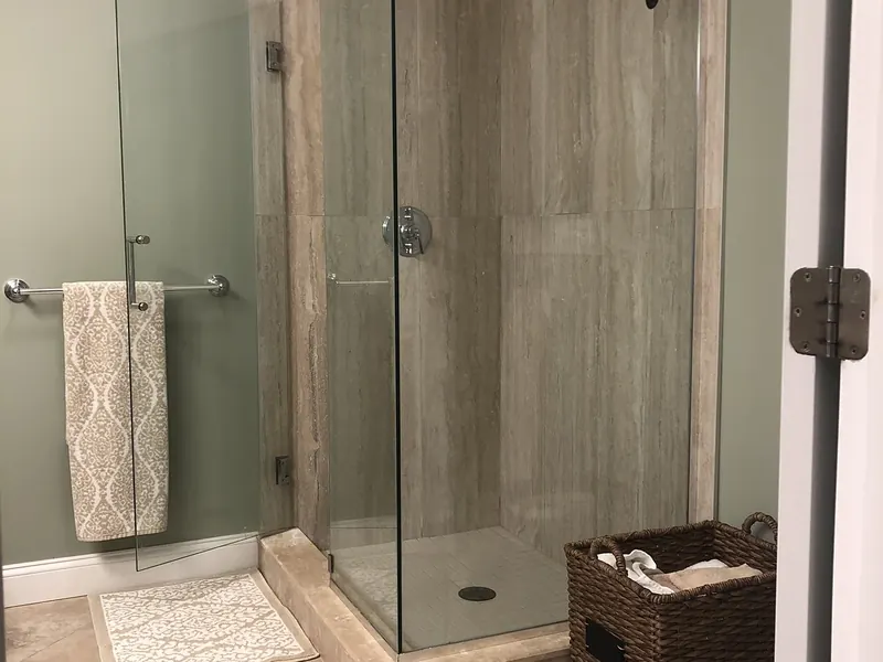 Corner frameless glass shower enclosure with travertine tile, rain shower head, and sage-green walls installed by A.J. LeBlanc Heating in a Southern NH home
