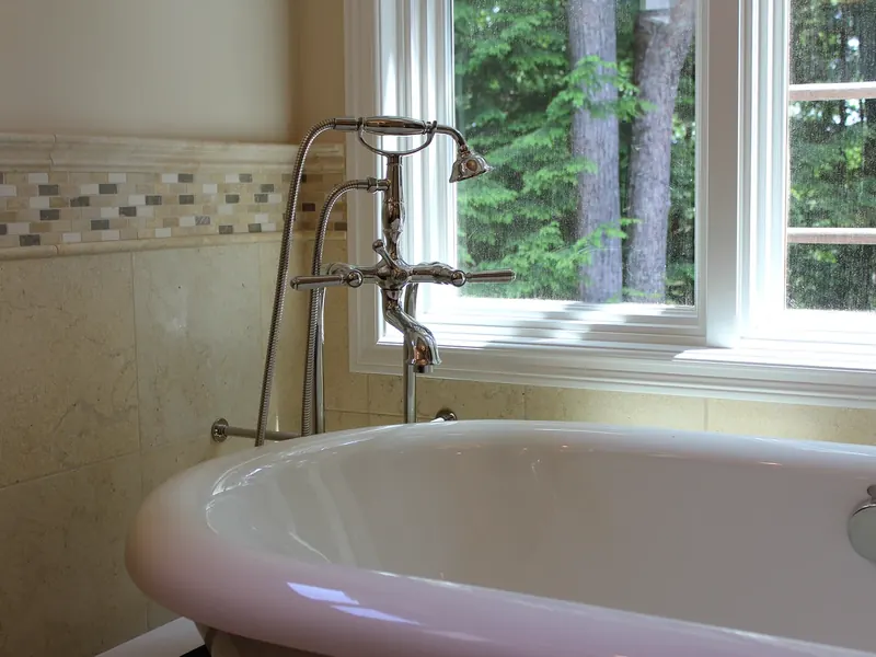 Freestanding bathtub with chrome telephone-style tub filler and handheld wand beside a window overlooking the woods, with travertine tile wall and glass mosaic accent band by A.J. LeBlanc Heating in a Southern NH home