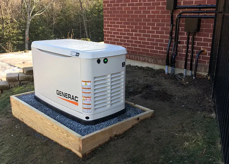17kW Generac whole-home standby generator on a wood-framed gravel pad against a red brick Southern NH home, installed by A.J. LeBlanc Heating