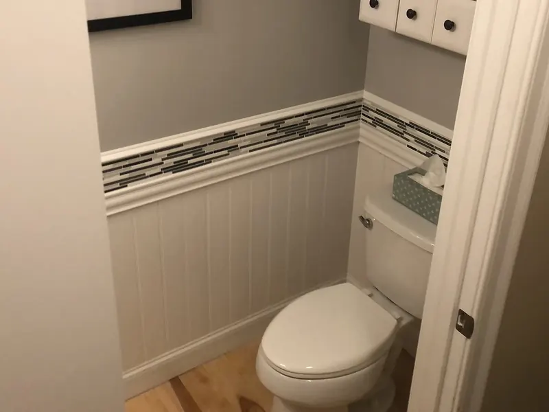 Half-bath toilet beside white wall cabinet with glass mosaic tile accent band, white beadboard wainscoting, and framed art by A.J. LeBlanc Heating in a Southern NH home