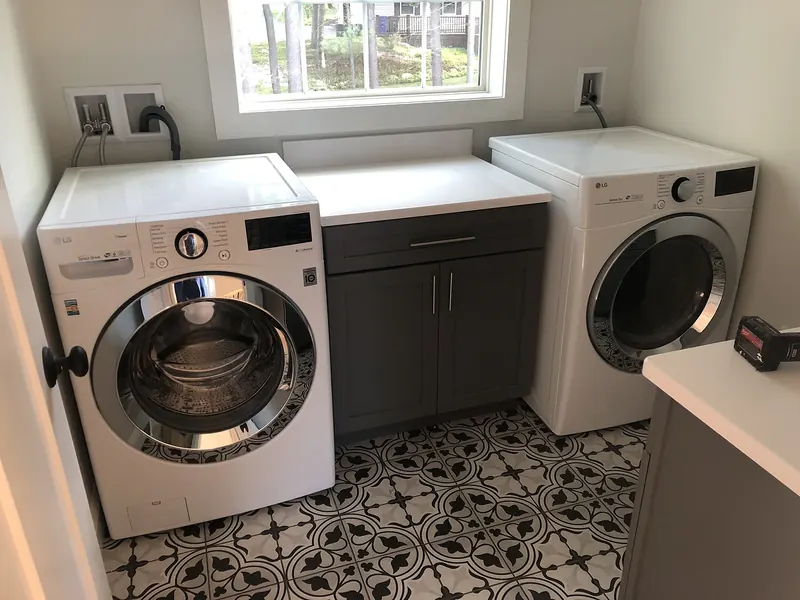 LG front-load washer and dryer side-by-side in a laundry room with patterned tile floor, gray cabinet, and window installed by A.J. LeBlanc Heating in a Southern NH home