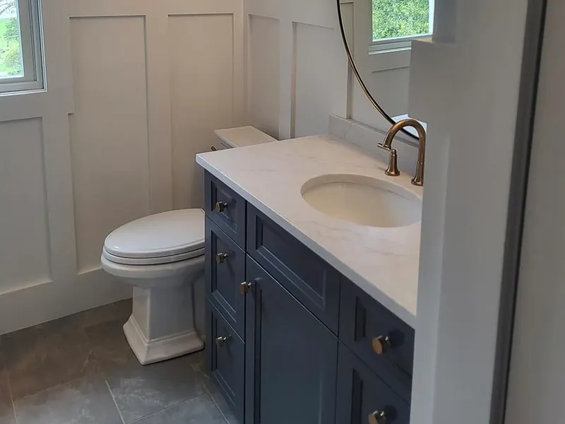 Master bathroom with round brass mirror, brass faucet, marble counter, navy vanity, and white wainscoting installed by A.J. LeBlanc Heating in a Southern NH home