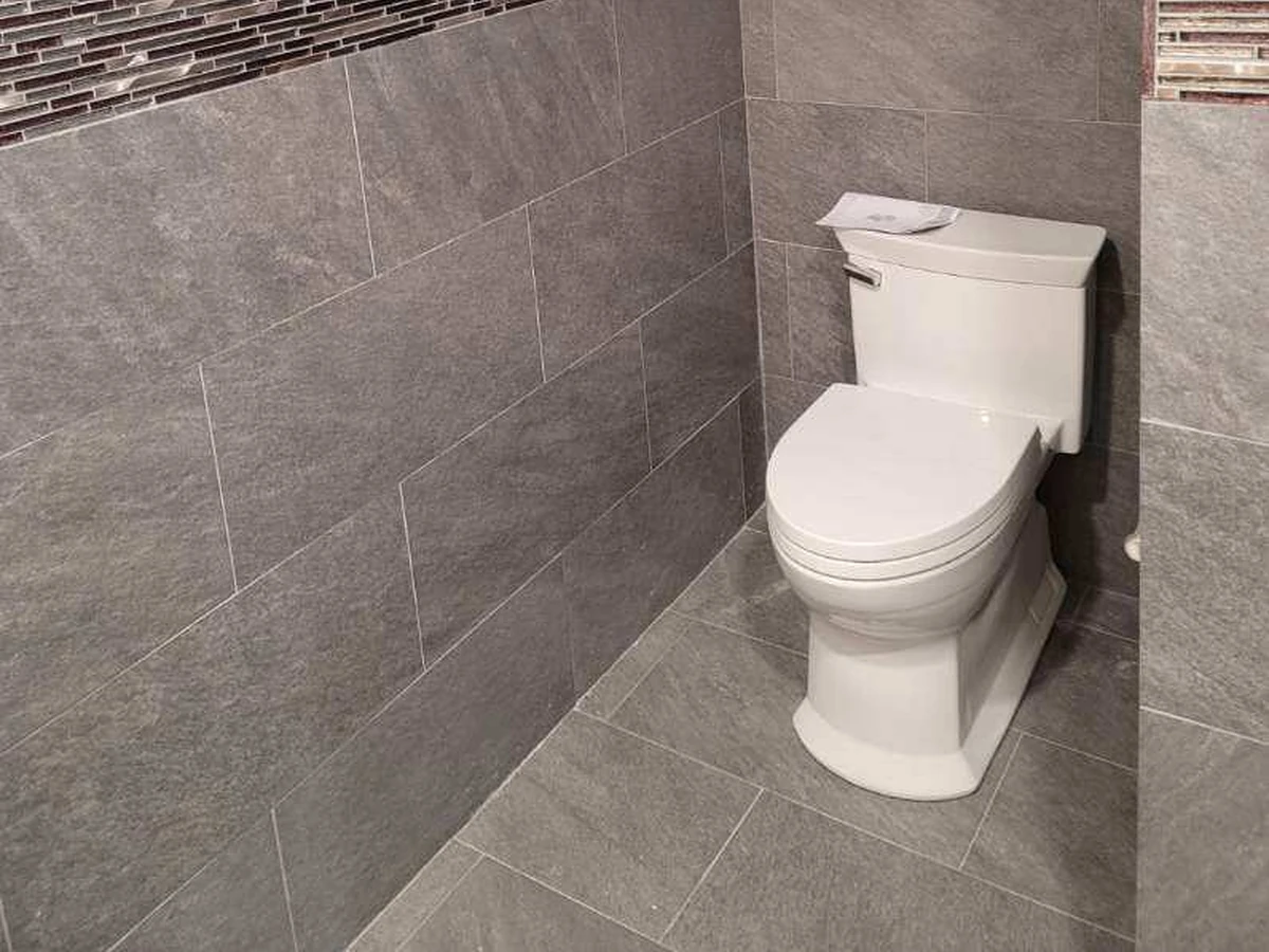 Modern skirted one-piece toilet installed in a designer bathroom with large gray porcelain tile, glass mosaic accent band, and an octagonal window by A.J. LeBlanc Heating in Southern NH