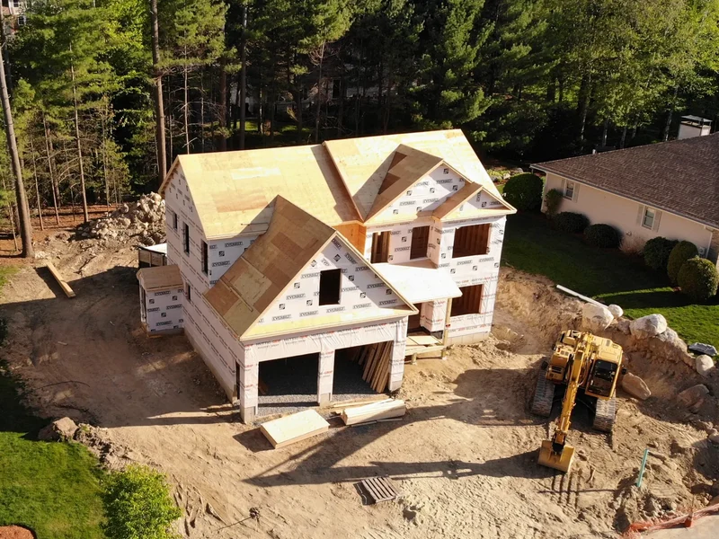 Aerial view of a two-story home under construction with sheathing in progress and an excavator on site