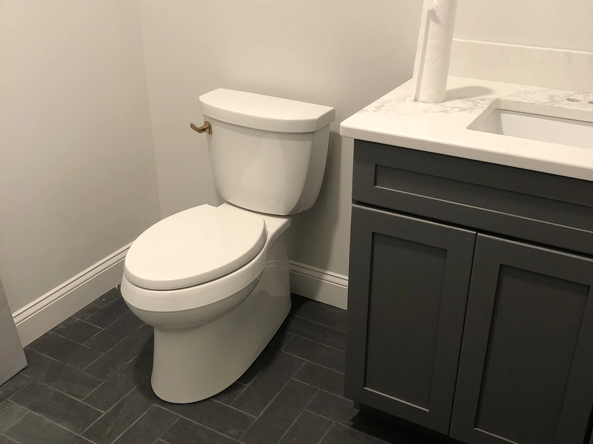 New construction toilet installation with gray shaker vanity, white quartz counter, and herringbone gray tile floor by A.J. LeBlanc Heating in Southern NH