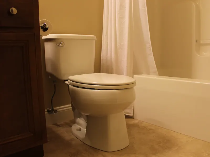 Close-up of a white two-piece toilet installation beside a brown wood vanity cabinet and a tub-shower combo with a white shower curtain installed by A.J. LeBlanc Heating in a Southern NH home