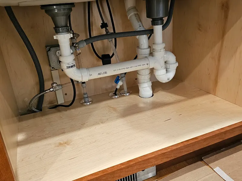 Under-sink plumbing replacement under a double-bowl kitchen sink with chrome basket strainers, white PVC continuous waste plumbing, dishwasher branch, and fresh supply shut-off valves by A.J. LeBlanc Heating in a Southern NH kitchen